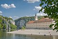 Kloster Weltenburg am Donaudurchbruch Flussufer mit Kiesstrand, altes Gebäude mit Turm und bewaldete Felsen unter blauem Himmel mit Wolken