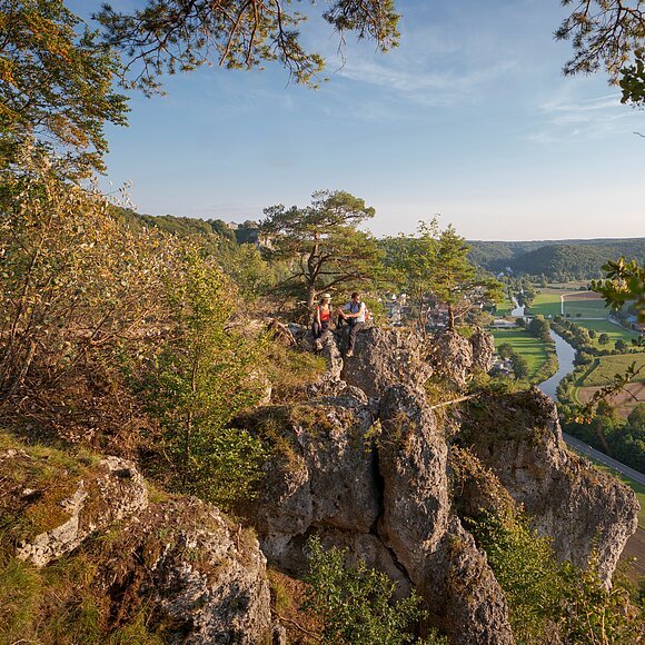 Ein Wanderpärchen, etwas entfernt, steht links auf einem Felsen an der Arnsberger Leite, die gut zu sehen ist. Dahinter erstreckt sich der Blick ins Tal. Der Himmel erstrahlt wolkenlos.