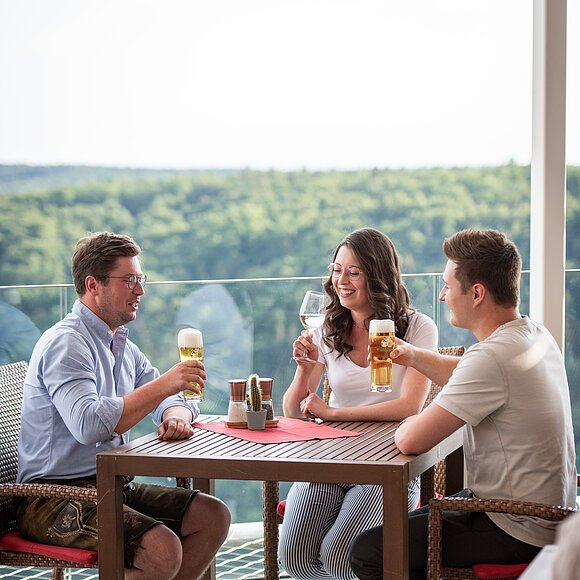 Zwei Männer und eine Frau, alle mit vollen Getränken in der Hand, prosten sich zu. Hinter ihnen hat man einen wunderschönen Blick ins Tal und auf die Wälder.