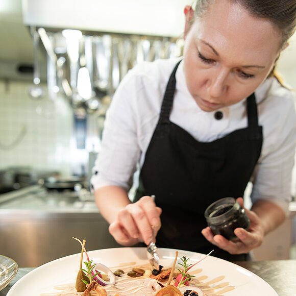 Eine Mitarbeiterin verleiht einer Vorspeise mit einem Pinsel und dunklen Soße den letzten Schliff. Hinter hier kann man Küchenutensilien erahnen.