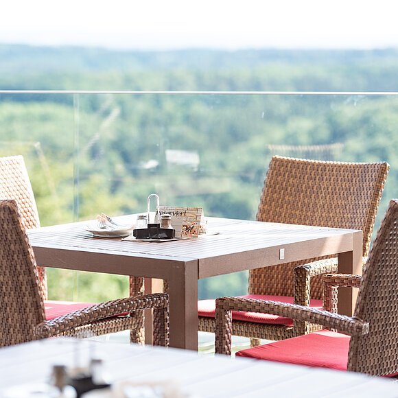 Ein Tisch mit vier Rattanstühlen mit roten Kissen. Dahinter eine Glasbrüstung und der wunderschöne Blick ins Tal und den grünen Wäldern.