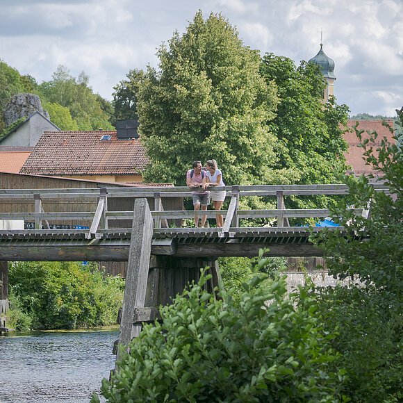 Wanderer auf der Brücke bei Essing An der Balustrade der Holzbrücke vor Essing lehnt sich ein Pärchen an und schaut flussaufwärts. Hinter ihnen sieht man einige Hausdächervon Essing umrahmt von Bäumen und deren Blätterdach.
