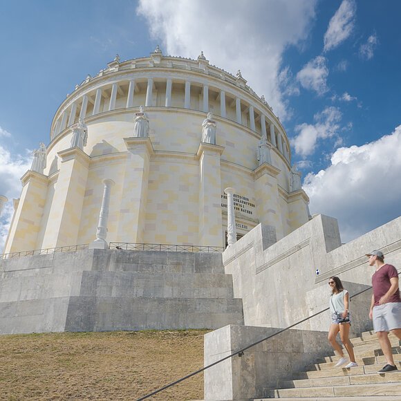 Befreiungshalle Kelheim Zwei Frauen und ein Mann, in sommerlicher Kleidung, steigen die Treppen der Befreiungshalle hinunter und werfen einen letzten Blick zu ihr hinauf. Der Himmel strahlt blau und ein paar Wolken sind zu sehen.