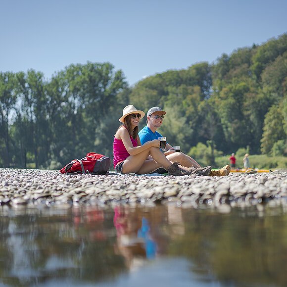 Ein Pärchen mit Hüten sitzt am Kiesstrand des Kloster Weltenburg. Vor ihnen fließt die Donau vorbei und im Hintergrund ist der Wald gegenüber zu sehen.