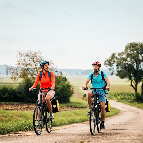 Eine Frau und ein Mann radeln auf dem Altmühltal-Radweg bei Dittenheim. Beide tragen einen Fahrradhelm und einen Rucksack. Der Radweg wird von Gras und grünen Bäumen gesäumt.