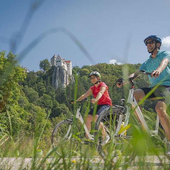 Zwei Radler, eine Frau mit weißem Helm und roten Shirt und ein Mann, vor ihr, ebenfalls mit Helm und blauen Shirt sind auf dem Radweg unterwegs. Die Zwei radeln und hinter ihnen ragt die Burg Prunn, die umgeben von Wäldern auf einem Felsen. Der Himmel scheint blau, nur ein paar Wolken verirren sich.