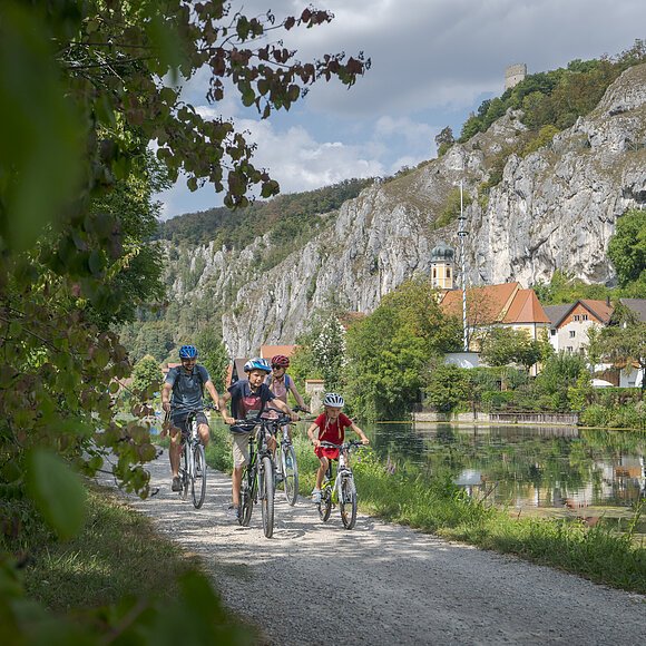 Eine vierköpfige Familie radelt entlang des Ludwig-Donau-Main-Kanal. Links ist der Kanal und der Ort Essing zu sehen. Dahinter erheben sich Felswände.