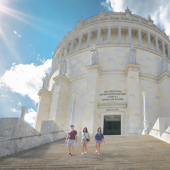 Zwei Frauen und ein Mann, in sommerlicher Kleidung, steigen die Treppen der Befreiungshalle hinunter. Der imposante Rundbau mit dessen Eingang ragt hinter ihnen auf. Der Himmel strahlt blau und ein paar Wolken sind zu sehen.