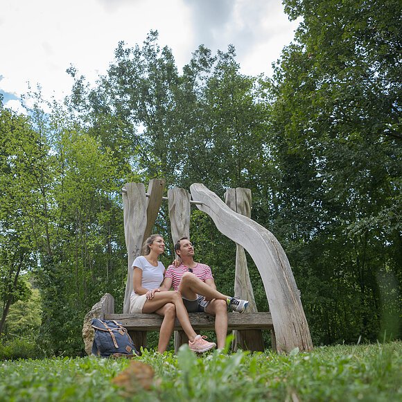 Ein Wanderpärchen hockt auf einer Bank entlang des Kunstweges in Essing. Hinter ihnen steht ein steinernes Kunstwerk. Die Bäume ringsherum sind im satten dunkelgrün. Der Himmel blitzt ein wenig durch.