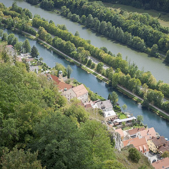 Wir blicken von oben auf Essing am Main-Donau-Kanal. Die Ortschaft ist von Wald, Felsen und auf der anderen Seite vom Kanal umgeben.