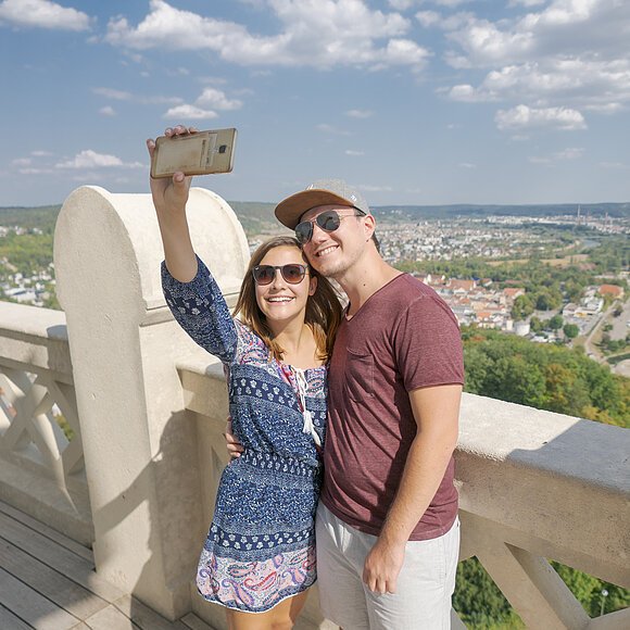 Ein Pärchen steht auf dem oberen Umgang, der um die Befreiungshalle führt. Die Frau hält ihr Smartphone in die Höhe und macht ein Selfie von sich und ihrem Freund, im Hintergrund Kelheim und der blaue, leicht bewölkte Himmel.