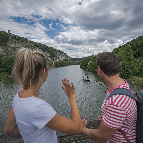 Eine Frau mit weißen Shirt und ein Mann mit Rucksack stehen mit dem Blick auf den Main-Donau-Kanal gerichtet auf der Holzbrücke am Geländer. Der Himmel ist sehr bedeckt. Die beiden Ufer erstrahlen im dunklen grün.