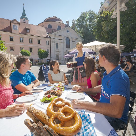 Fünf Personen sitzen an einem Tisch im Freien, eine Kellnerin bringt Bier, Brezeln und Brot liegen auf dem Tisch.