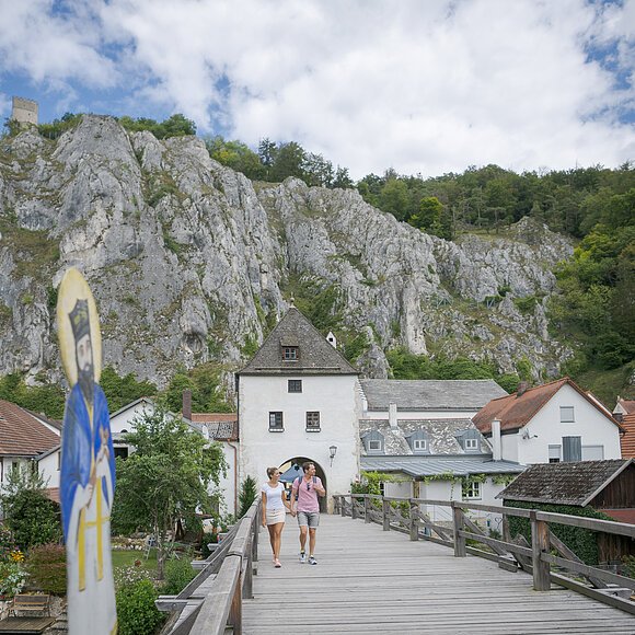 Paar geht auf einer Holzbrücke zu einem historischen Torhaus vor Felsen und Häusern unter bewölktem Himmel.