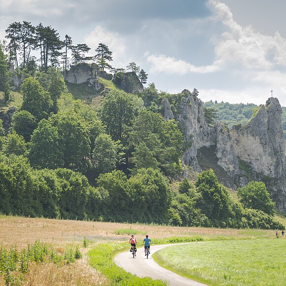 Zwei Radfahrer auf einem Weg vor bewaldeten Felsen mit einem Kreuz auf dem höchsten Felsen.