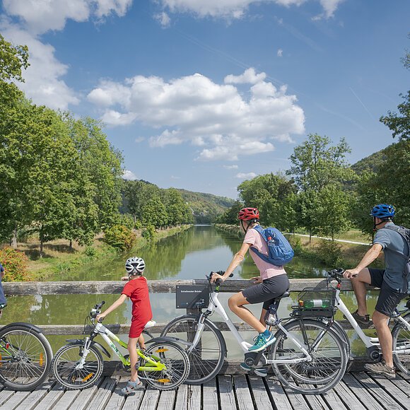 Vier Personen mit Fahrrädern auf einer Holzbrücke mit Blick auf einen Fluss und grüne Landschaft.