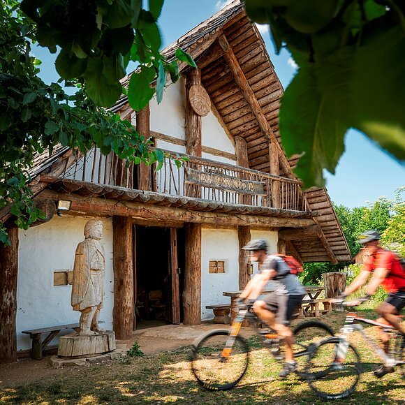Holzhaus mit Statue davor und zwei Radfahrern, umgeben von Bäumen bei sonnigem Wetter