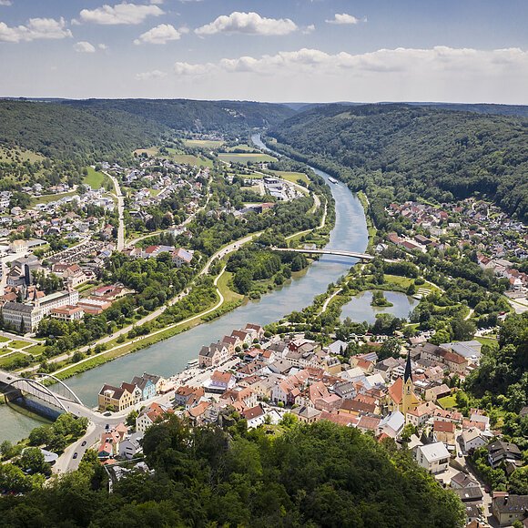 Der Blick auf Riedenburg von oben. Mitten durch die Stadt verläuft der Main-Donau-Kanal. Umrandet wird sie von grünen Wäldern. Der Himmel ist blau und einige Wolken sind zu sehen.