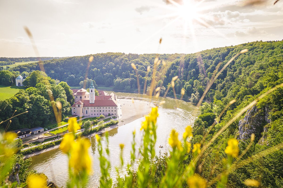 Fluss mit Schloss am Ufer, umgeben von Wald und gelben Blumen im Vordergrund bei Sonnenschein