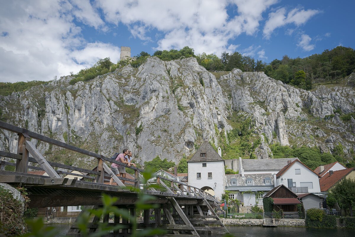 Wanderer in Essing An der Balustrade der Holzbrücke vor Essing lehnt sich ein Pärchen an und schaut flussabwärts. Hinter ihnen sieht man einige Häuser von Essing, dahinter die aufragenden Felswände.