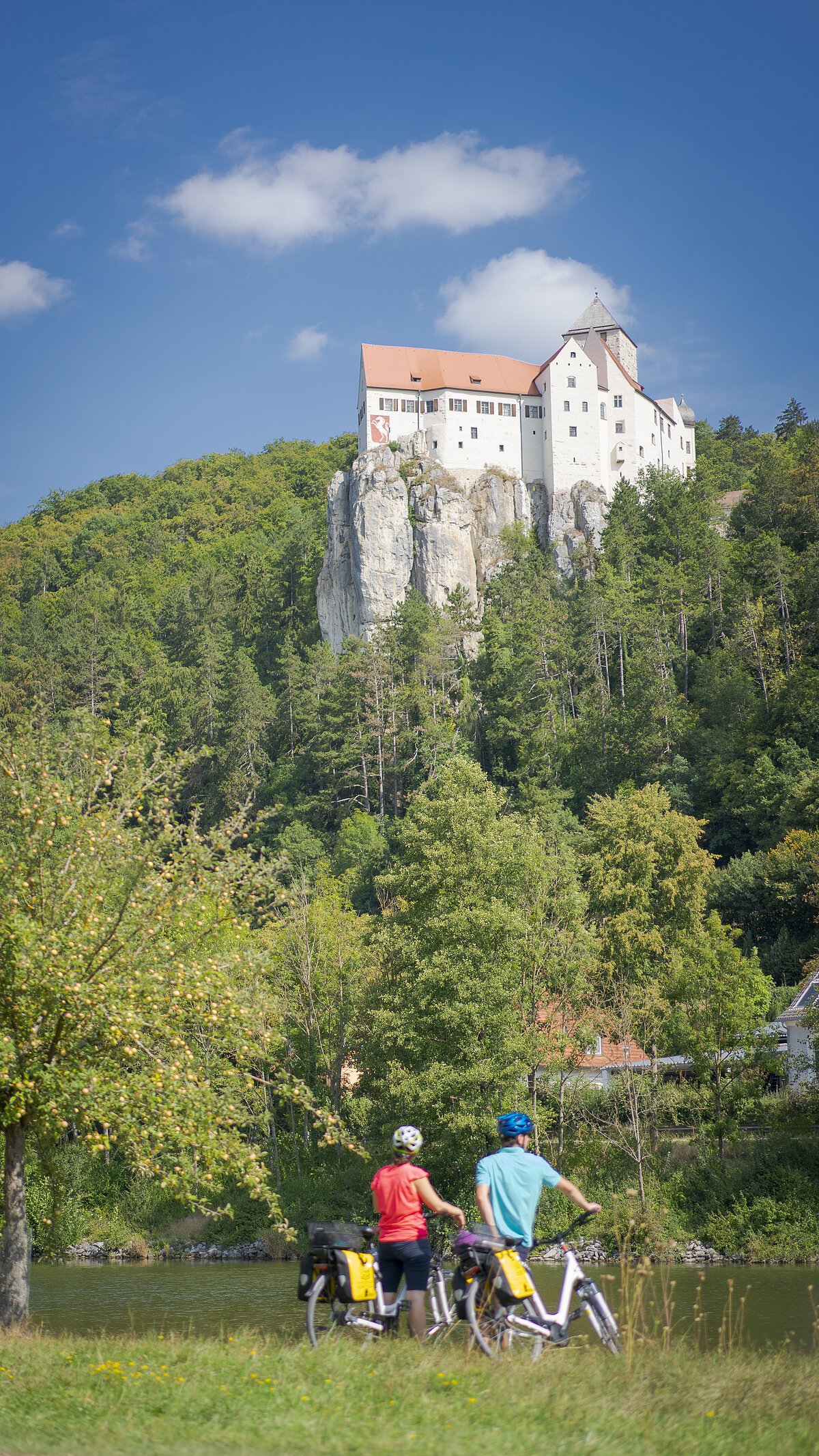 Zwei Radfahrer stehen an einem Fluss, im Hintergrund ein weißes Schloss auf einem Felsen und bewaldete Hügel.