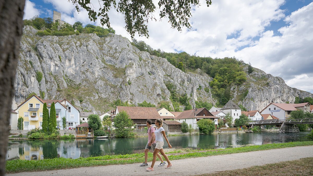 Ein Pärchen spaziert entlang des Ludwig-Donau-Main-Kanals. Im Hintergrund ist der Ort Essing und seine Felsen zu sehen.
