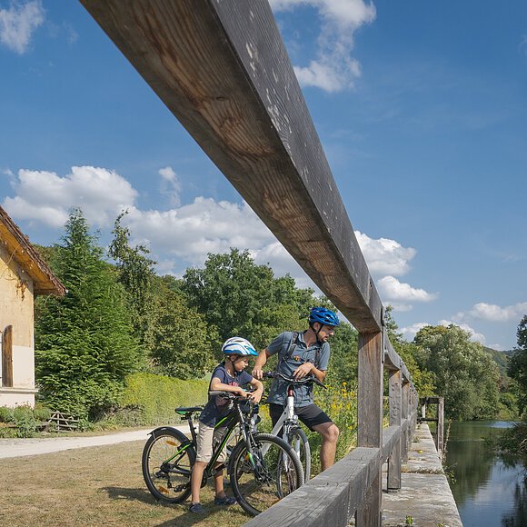 Radlerfamilie macht halt am Schleusenhäuschen Mühlbauch Familie mit vier Personen und Fahrrädern neben einem alten Haus und einem Fluss unter blauem Himmel.