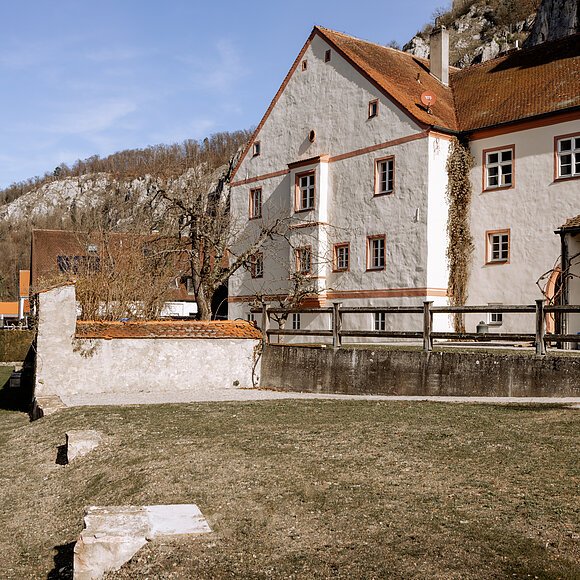 MEMU Essing Außenansicht Großes, weißes Gebäude mit roten Dächern, umgeben von Bäumen und Wiese, im Hintergrund Hügel und blauer Himmel.